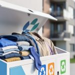 Overfilled textile recycling drop-off bin with assorted clothing, photographed in a clean outdoor Canadian setting under soft daylight.